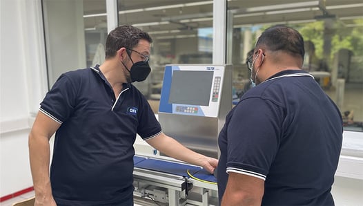 Two CASSEL Inspection technicians stand looking at a checkweigher in a lab setting.