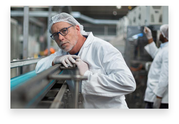 A caucasian middle-aged man with a hair net and white lab coat leaning on a stainless steel machine looking at the camera.