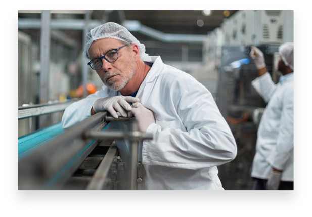 Ein kaukasischer Mann mittleren Alters mit Haarnetz und weißem Laborkittel lehnt an einer Edelstahlmaschine und schaut in die Kamera.